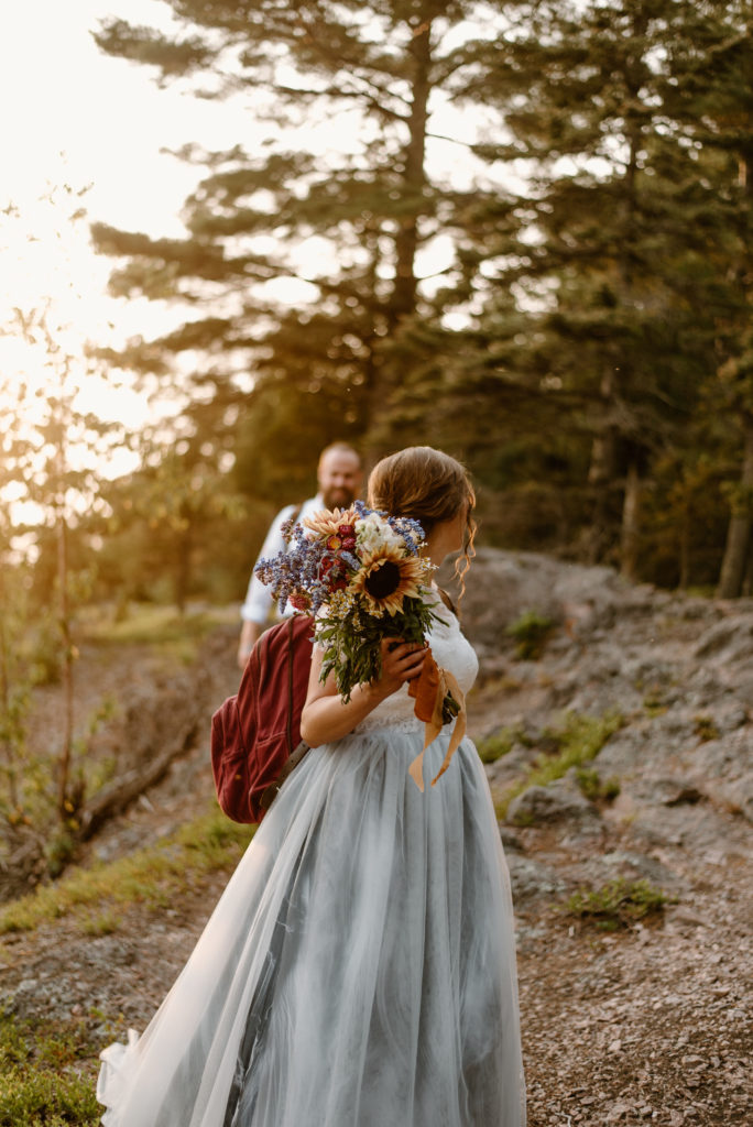 A bride looking back at her groom, with her wildflower bouquet in focus.  The couple is hiking along the north shore of Lake Superior in the Superior National Forest during golden hour.